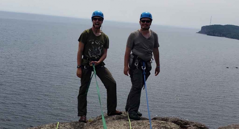 two people in rock climbing gear smile at the camera while standing on a cliff high above a body of water